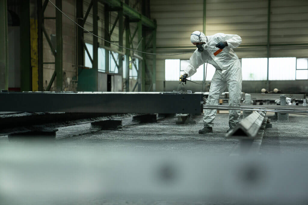 Eine Person in Schutzkleidung und mit Atemschutzmaske beschichtet mit einer Spritzpistole einen großen Metallträger in einer industriellen Halle mit hohen Decken und grünen Stützbalken.