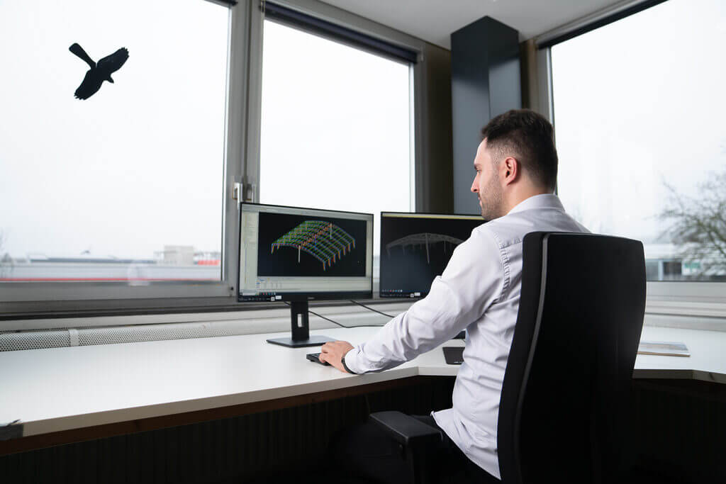 Ein Mann sitzt an einem Schreibtisch und arbeitet an einem Computer mit zwei Monitoren, auf denen Architektur- oder Bauplanungssoftware läuft. Hinter ihm befinden sich große Fenster, und draußen fliegt ein Vogel.