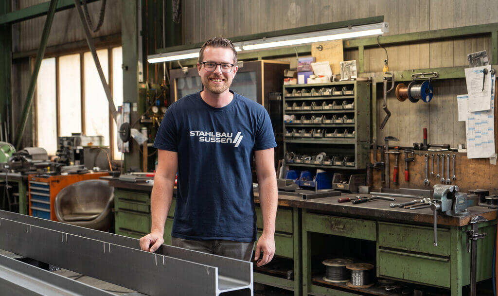 Ein Mann mit Brille und einem blauen T-Shirt mit der Aufschrift „Stahlbau Sussen“ steht lächelnd in einer Werkstatt, im Hintergrund sind grüne Werkbänke, Werkzeuge, Regale und Industrieanlagen zu sehen.